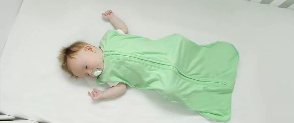 A baby sleeps peacefully in a light green sleep sack on a white crib mattress.