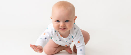 The image shows a baby in a white onesie with colorful polka dots, in a crawling position, looking forward with a smile, appearing happy and engaged.