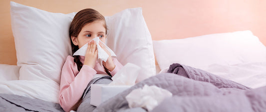 A young girl with a cold sits in bed, blowing her nose with a tissue, surrounded by more tissues and a cosy blanket.