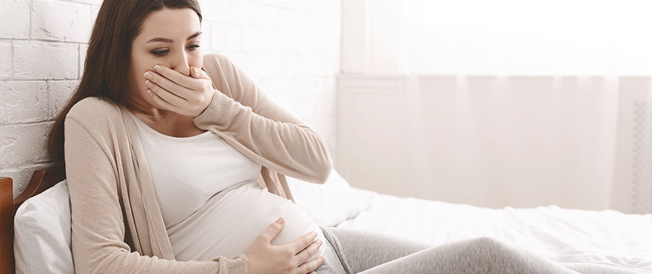 A pregnant woman sitting on a bed, covering her mouth, feeling nauseous.