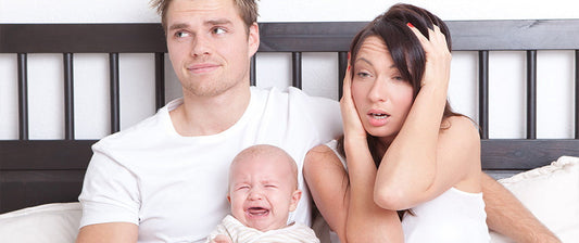 A tired couple looks exhausted in bed while their baby cries beside them.