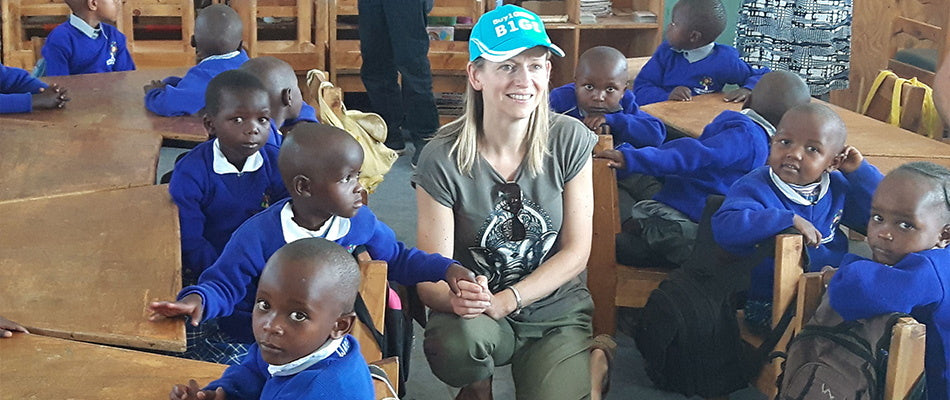 A woman sits among a group of young students wearing school uniforms in a classroom, sharing a moment of cultural exchange and education.
