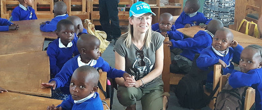A woman sits among a group of young students wearing school uniforms in a classroom, sharing a moment of cultural exchange and education.
