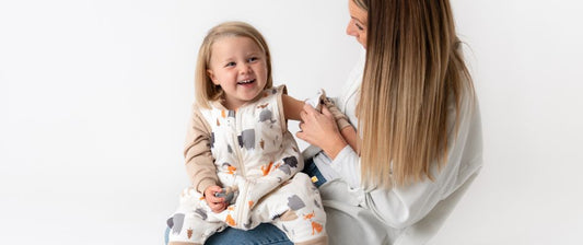  A joyful toddler in a patterned outfit laughs while sitting on her mother's lap, who is smiling and engaging warmly with her child against a clean, white background.