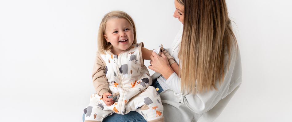  A joyful toddler in a patterned outfit laughs while sitting on her mother's lap, who is smiling and engaging warmly with her child against a clean, white background.