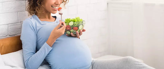 A smiling pregnant woman sits on a bed, eating a salad from a bowl.