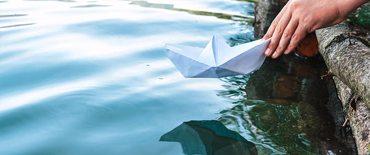 A child's hand launches a paper boat into tranquil blue water from a wooden edge.