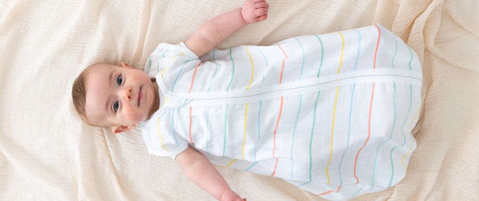 A baby in a striped baby sleeping bag lies on a cream-colored blanket.