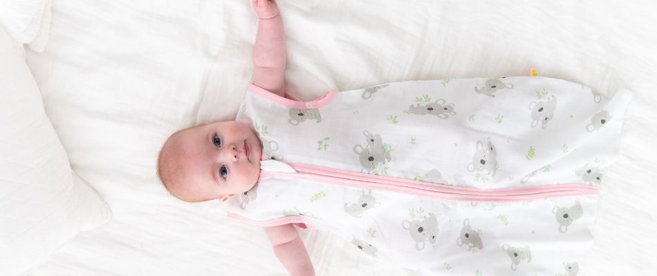 A baby in a sleep sack with koala print, lying on a white bed.