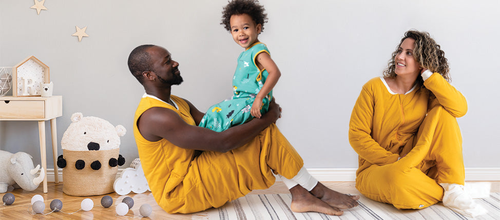 A joyful family scene in a nursery: a father lifting his smiling toddler, with the mother sitting nearby, all dressed in matching yellow outfits.