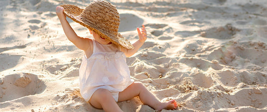 a young child sitting on a sandy beach. The child is wearing a light, airy dress and playfully holding a large straw hat over their head.