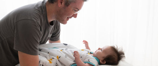 A father leans over affectionately towards his baby who is lying on a bed, both engaging in a delightful interaction.