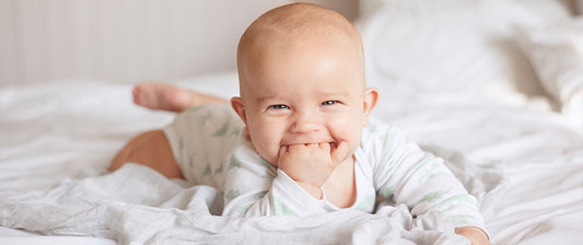  A cheerful baby lies on a white bed, smiling and sucking on their hand, capturing a delightful moment of early development.