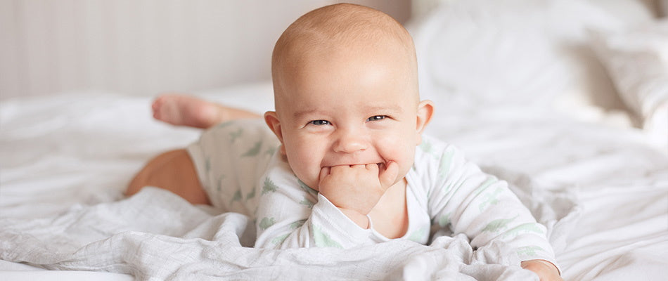  A cheerful baby lies on a white bed, smiling and sucking on their hand, capturing a delightful moment of early development.