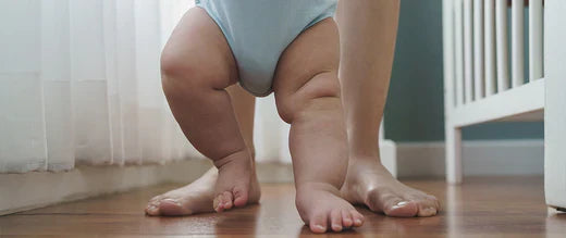  Close-up of a baby's chubby legs taking first steps, guided by an adult's feet nearby.