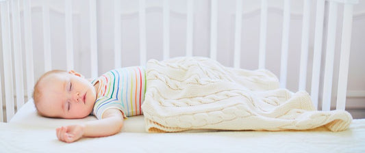 a peaceful baby asleep in a crib, covered by a cosy, knitted blanket. The baby is lying on their back, wearing a colourful striped onesie. 