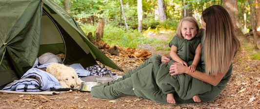  A mother and her young child, both dressed in green, share a joyful moment outside a tent during a camping trip in the forest. A golden retriever rests inside the tent