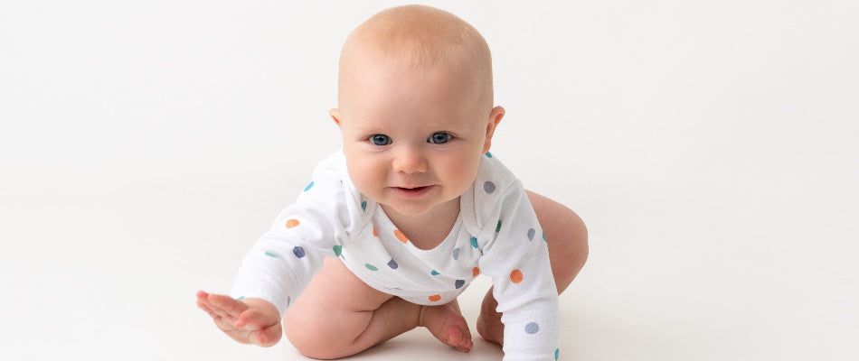 The image shows a baby in a white onesie with colorful polka dots, in a crawling position, looking forward with a smile, appearing happy and engaged.