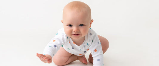 The image shows a baby in a white onesie with colorful polka dots, in a crawling position, looking forward with a smile, appearing happy and engaged.