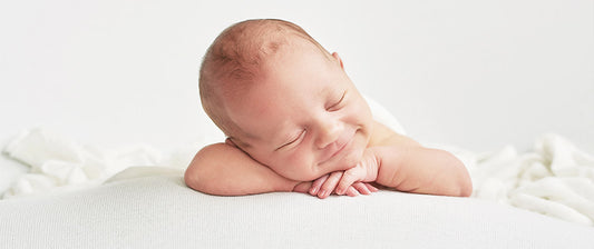 The baby is resting on their arms, creating a sweet and serene scene. The simple white backdrop and bedding emphasize purity and calmness.