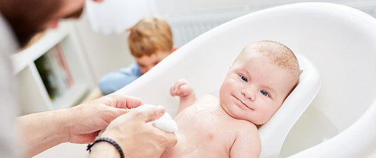  A smiling baby enjoys a bath in a white tub, watched over by a parent, while an older sibling plays in the background.
