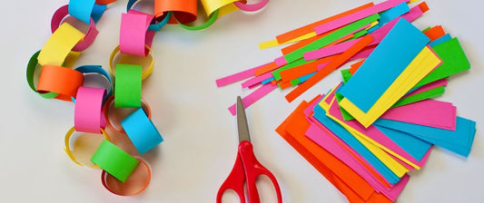 a colorful arrangement of craft supplies including paper chains made from brightly colored paper, alongside piles of cut and uncut paper strips in various colors