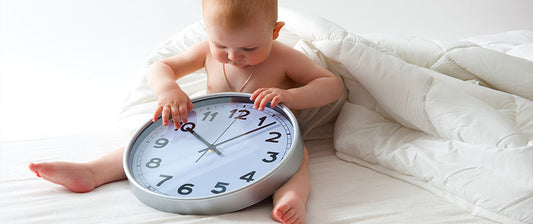 a baby sitting on a bed, closely examining a large analog clock. The setting appears to be a home environment with a simple and cozy backdrop, including a white duvet. 