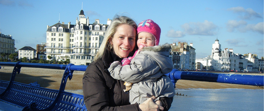  A woman holding a child smiles warmly at the camera against a picturesque seaside backdrop with historic buildings in the background, conveying a joyful and scenic moment outdoors.