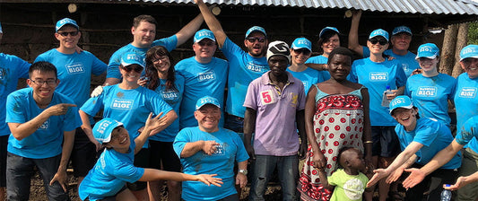  A group of volunteers in matching blue shirts pose happily with local residents in a rural setting, demonstrating a moment of community and collaboration during a service trip.