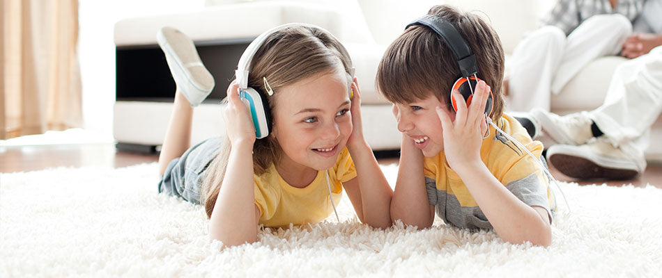 two young children, a girl and a boy, lying on a plush carpet and wearing headphones, seemingly enjoying music or an audio story together. 