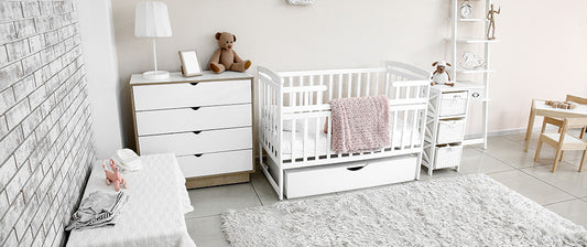 Nursery features a classic white crib with a teddy bear and a pink blanket, a matching white dresser, and a small chair with a table. The floor is covered by a plush grey area rug,