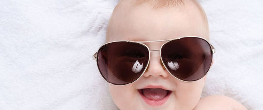  A cheerful baby wearing oversized sunglasses lies on a soft white blanket, embodying a playful approach to sun protection.