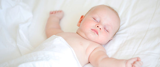 A baby sleeps peacefully on a white duvet, arms gently outstretched.