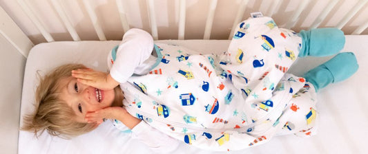 A cheerful toddler lies in a crib, smiling and playfully covering their face with hands, dressed in a colorful sleeping bag with feet.