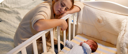 A weary mother rests her head on the railing of a crib, gazing affectionately at her newborn baby lying comfortably inside, capturing a tender yet exhausting moment of new parenthood.