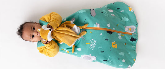 a baby lying on a flat surface, wrapped in a cozy, forest-themed baby sleeping bag. The sleeping bag features illustrations of woodland animals like bears and trees against a green background, with a yellow zipper running down the middle
