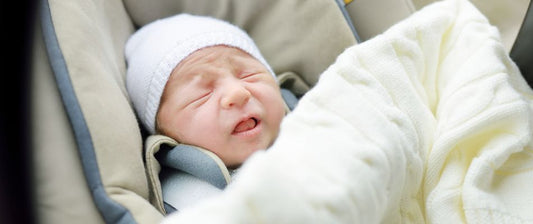  A newborn baby cries in a car seat, snugly wrapped and wearing a white hat, expressing discomfort or need.