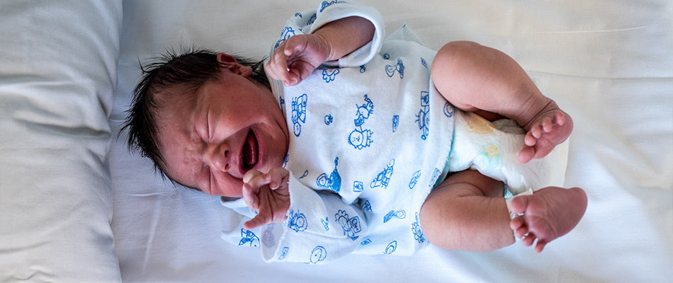 A newborn baby cries while lying on a white bed, wearing a onesie with a blue animal print, expressing early signs of discomfort or sleep issues.