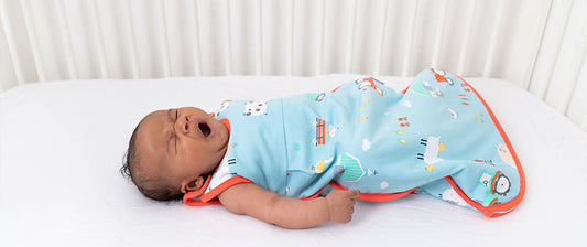 A yawning baby dressed in a colorful sleep sack lies on a white bed.