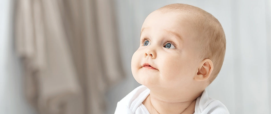 A curious baby with blue eyes looks up, dressed in a white outfit against a soft, neutral background.