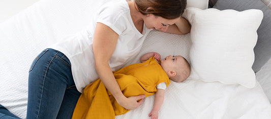  A mother tenderly looks at her baby, who is dressed in a yellow outfit, as they lie together on a white bed.