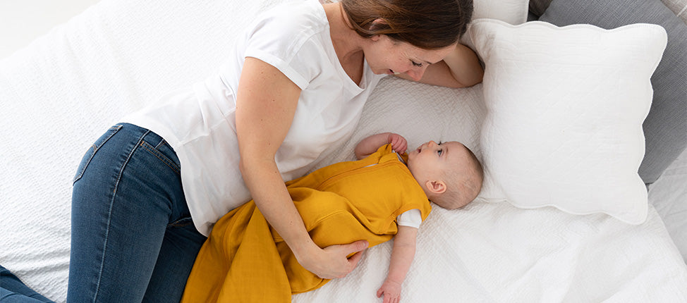 A mother tenderly looks at her baby, who is dressed in a yellow outfit, as they lie together on a white bed.