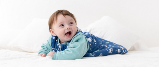 A joyful baby in a blue sleeping bag, lying on a bed, laughs delightedly.